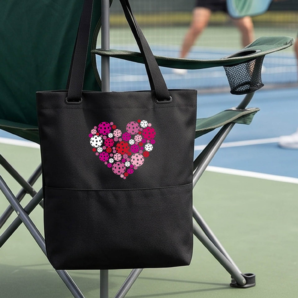 A black tote bag with a colorful heart-shaped floral pattern made of pickleballs  on the front, hanging from a folding chair on a pickleball court.