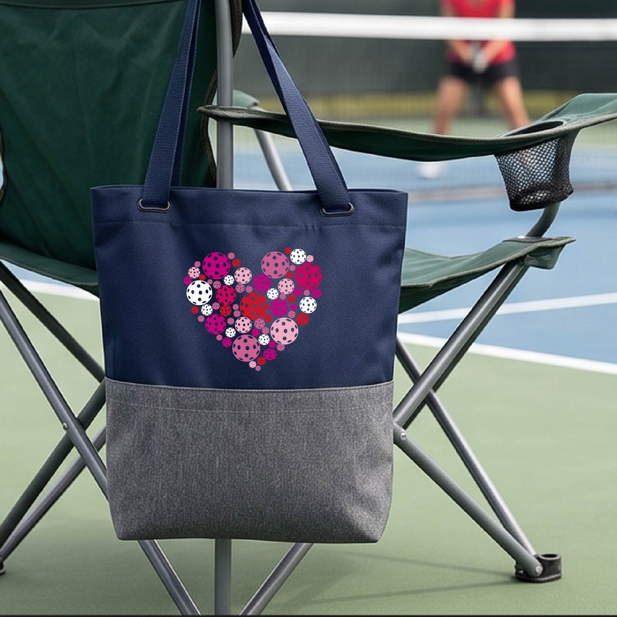 A blue and gray tote bag with a colorful heart-shaped floral pattern made of pickleballs  on the front, hanging from a folding chair on a pickleball court.