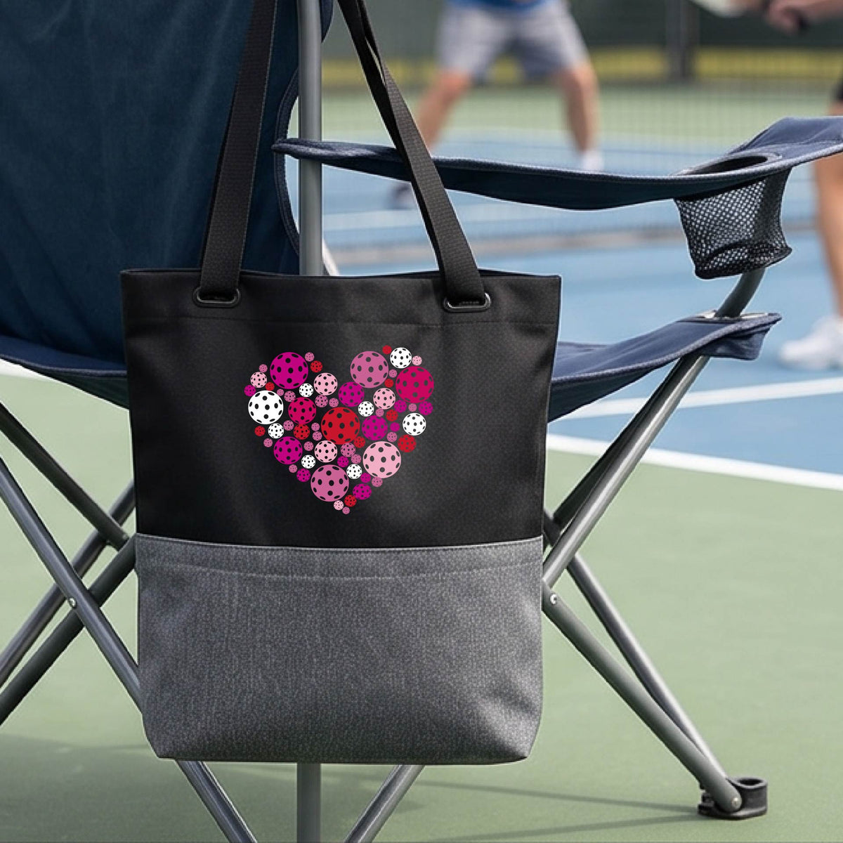 A black and gray tote bag with a colorful heart-shaped floral pattern made of pickleballs  on the front, hanging from a folding chair on a pickleball court.