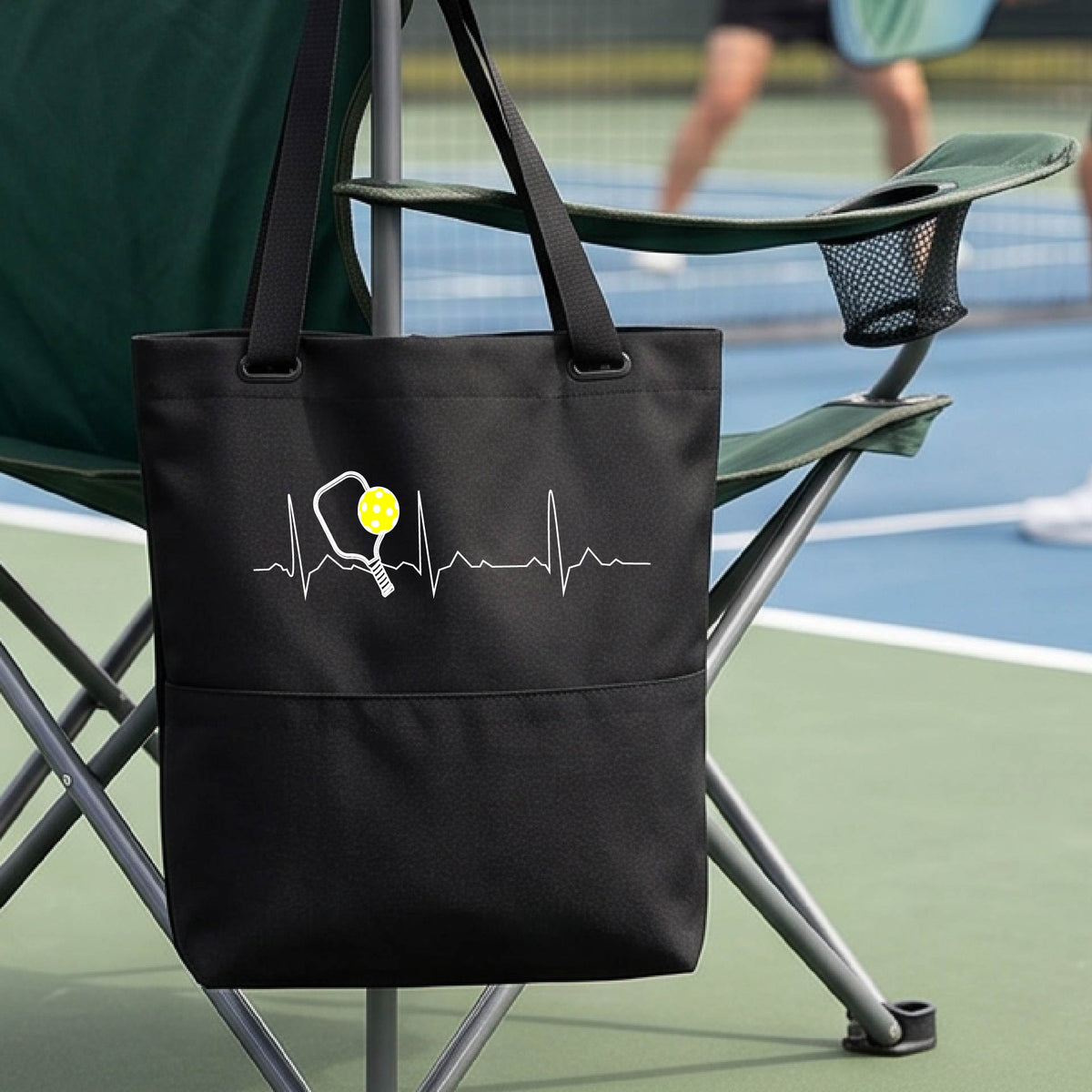 A close-up of a black tote bag sporting a white pickleball graphic of a heartbeat EKG including a pickleball paddle and a yellow pickleball across the check, in front of a pickleball court.