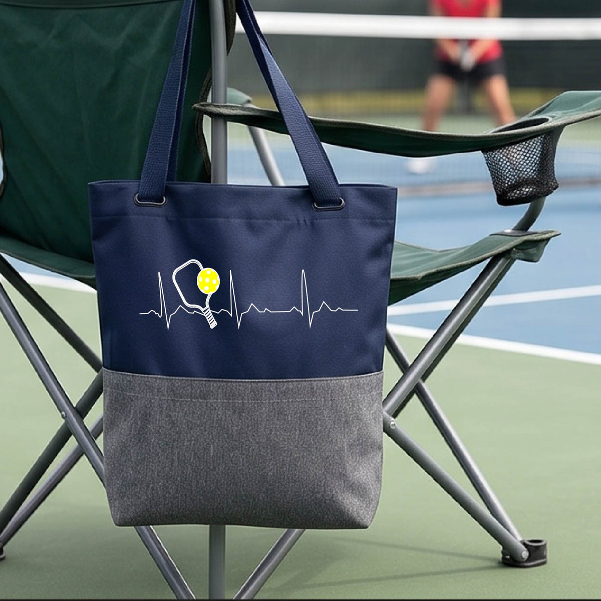 A close-up of a navy and gray tote bag sporting a white pickleball graphic of a heartbeat EKG including a pickleball paddle and a yellow pickleball across the check, in front of a pickleball court.