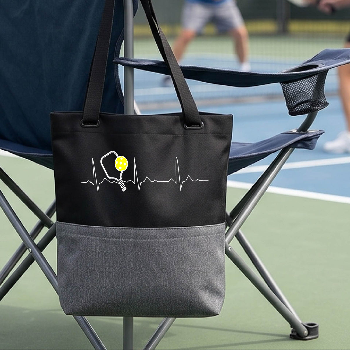 A close-up of a gray and black tote bag sporting a white pickleball graphic of a heartbeat EKG including a pickleball paddle and a yellow pickleball across the check, in front of a pickleball court.