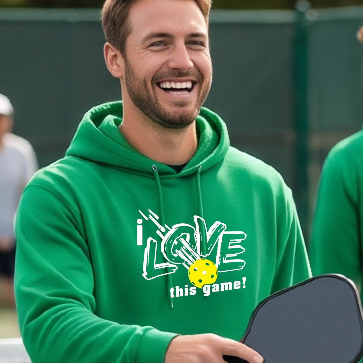 A smiling man wearing a bright green hoodie with a graphic that reads 'i LOVE this game!' and a yellow pickleball, photographed outdoors on a pickleball court.