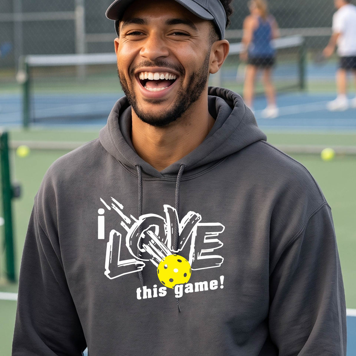 A smiling man wearing a dark gray hoodie with a graphic that reads 'i LOVE this game!' and a yellow pickleball, photographed outdoors on a pickleball court.