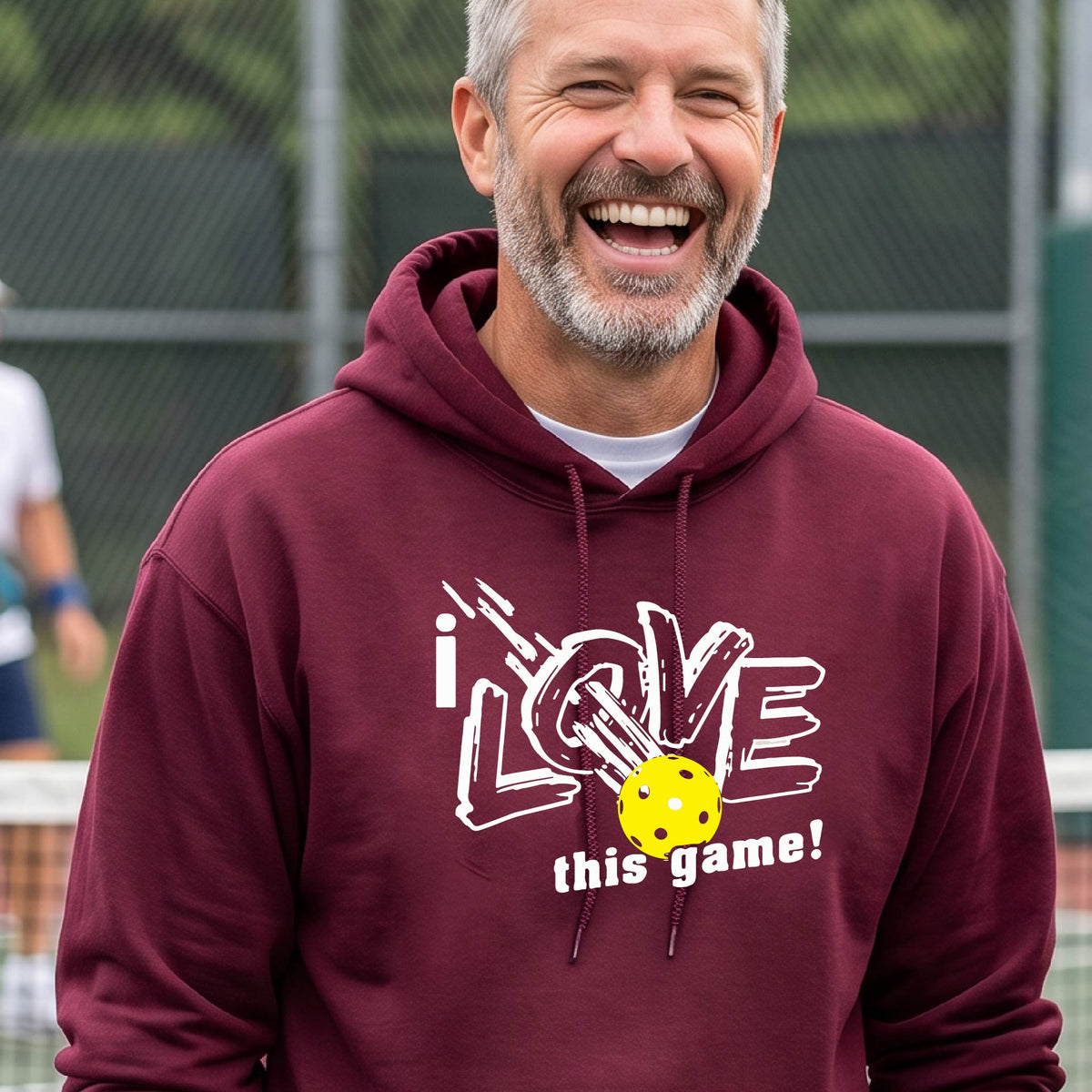 A smiling man wearing a maroon hoodie with a graphic that reads 'i LOVE this game!' and a yellow pickleball, photographed outdoors on a pickleball court.