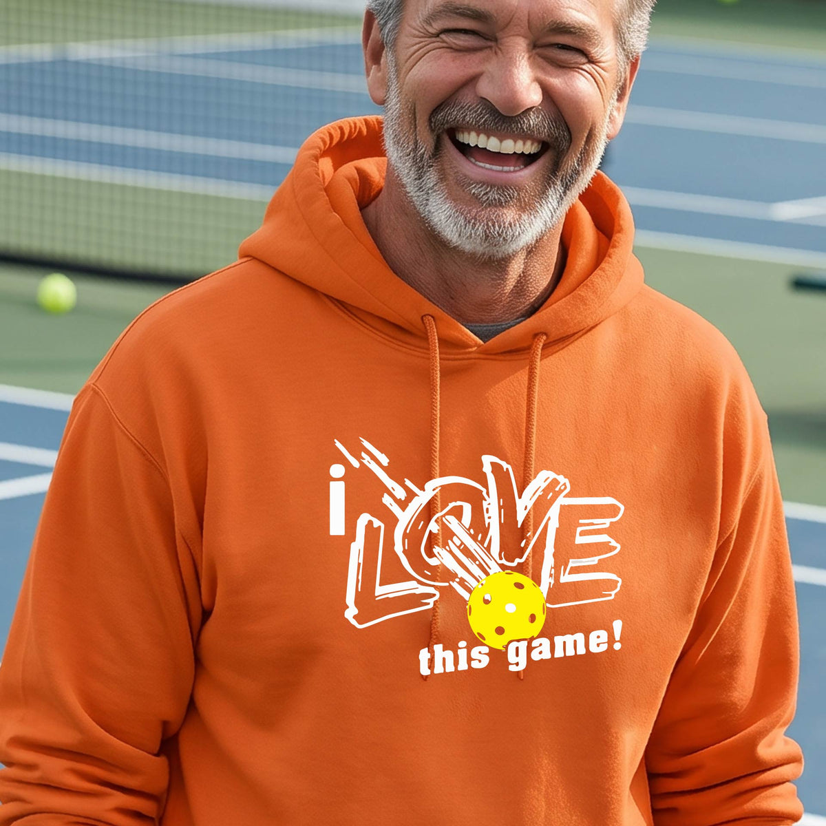 A smiling man wearing a bright orange hoodie with a graphic that reads 'i LOVE this game!' and a yellow pickleball, photographed outdoors on a pickleball court.