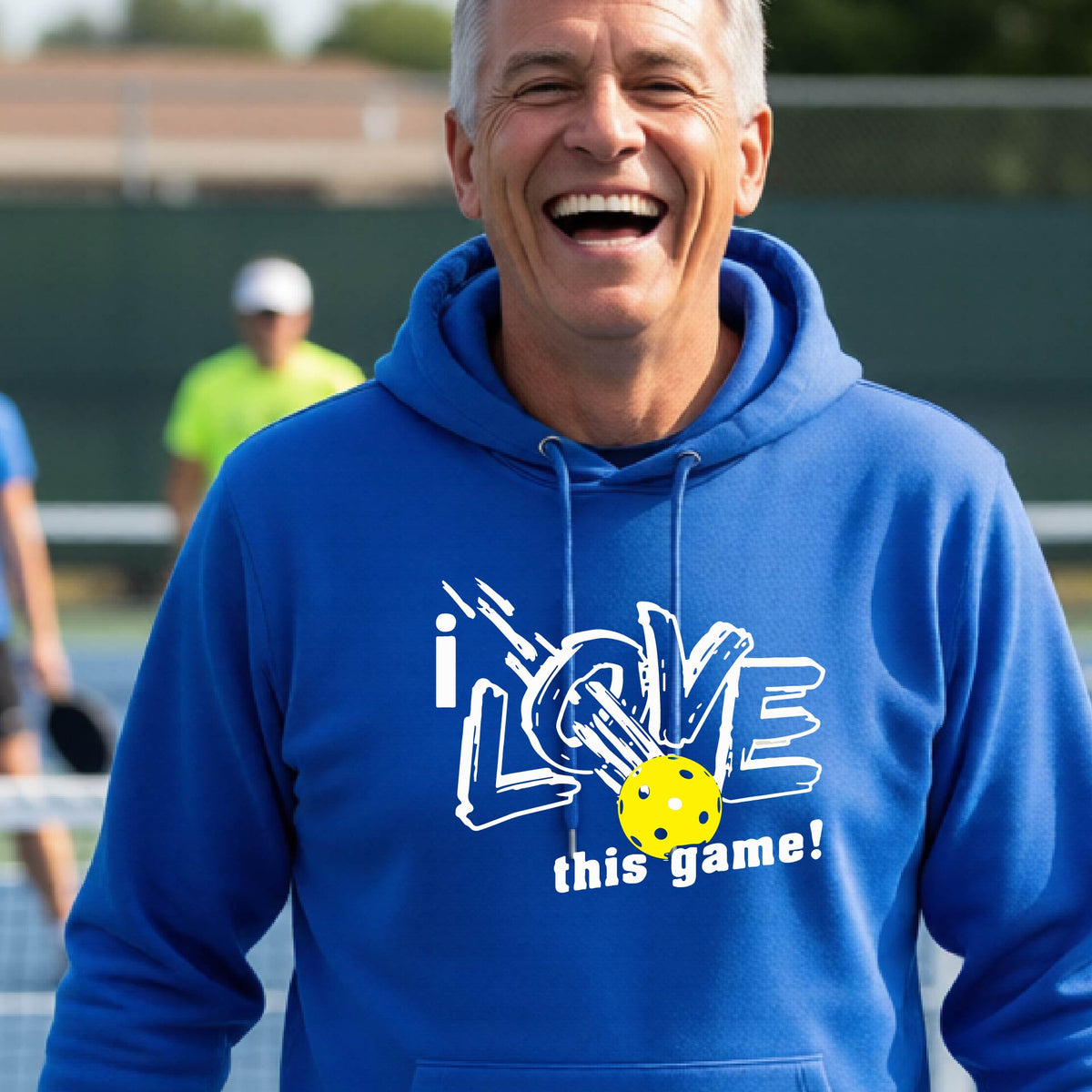 A smiling man wearing a bright blue hoodie with a graphic that reads 'i LOVE this game!' and a yellow pickleball, photographed outdoors on a pickleball court.