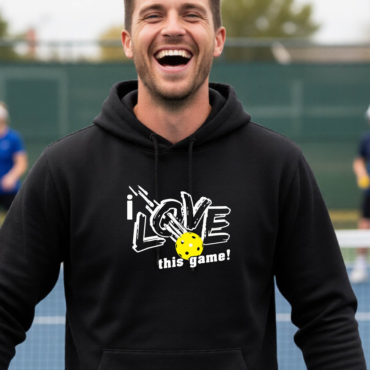 A smiling man wearing a black hoodie with a graphic that reads 'i LOVE this game!' and a yellow pickleball, photographed outdoors on a pickleball court.