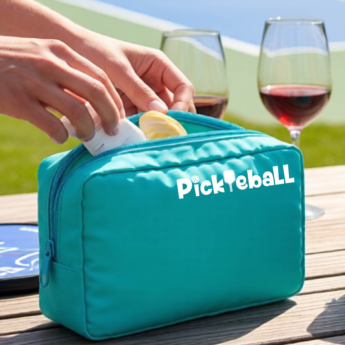 A turquoise cosmetic toiletry bag with a white graphic of the word 'Pickleball' and an intergrated pickleball paddle replacing the L. 