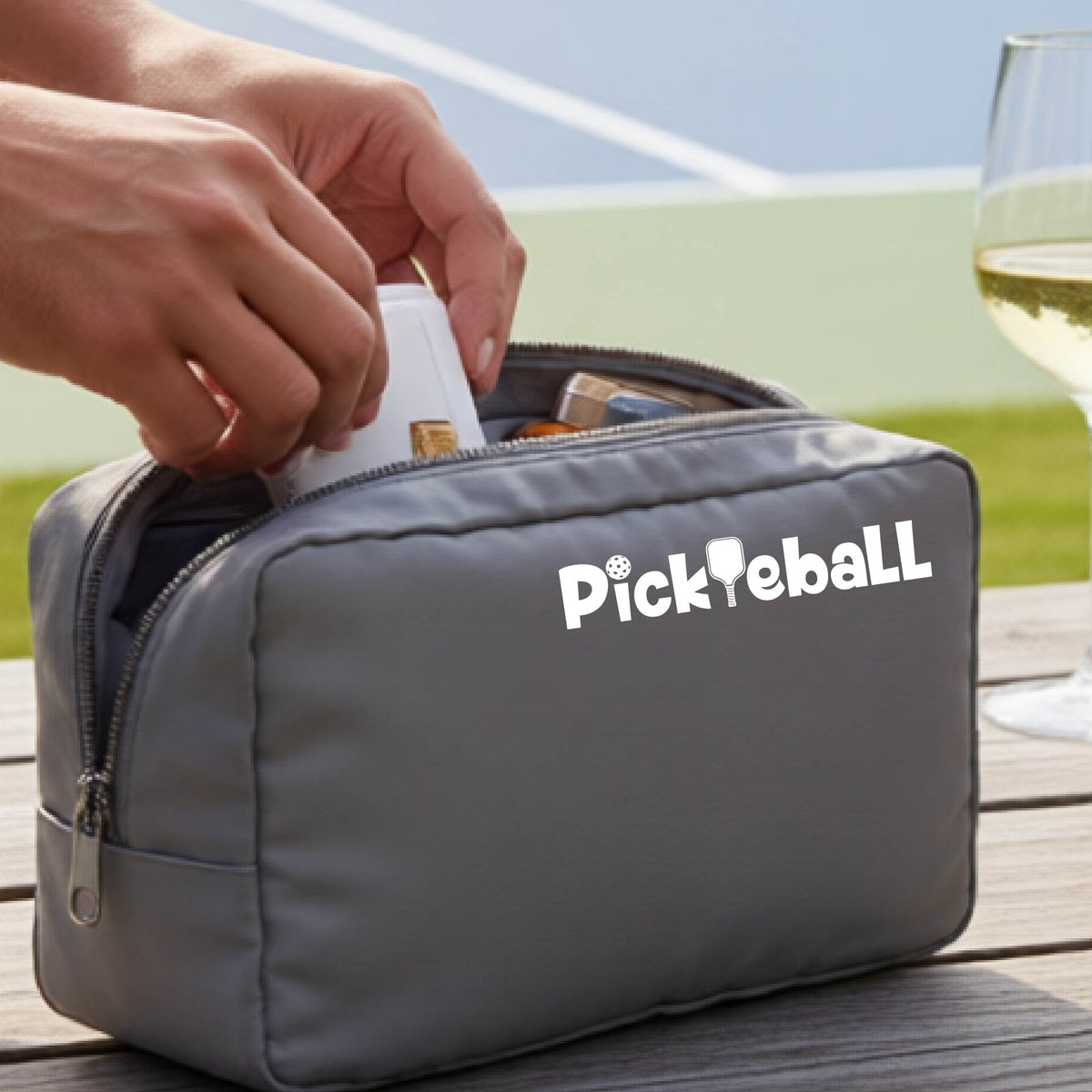 A gray cosmetic toiletry bag with a white graphic of the word 'Pickleball' and an intergrated pickleball paddle replacing the L. 