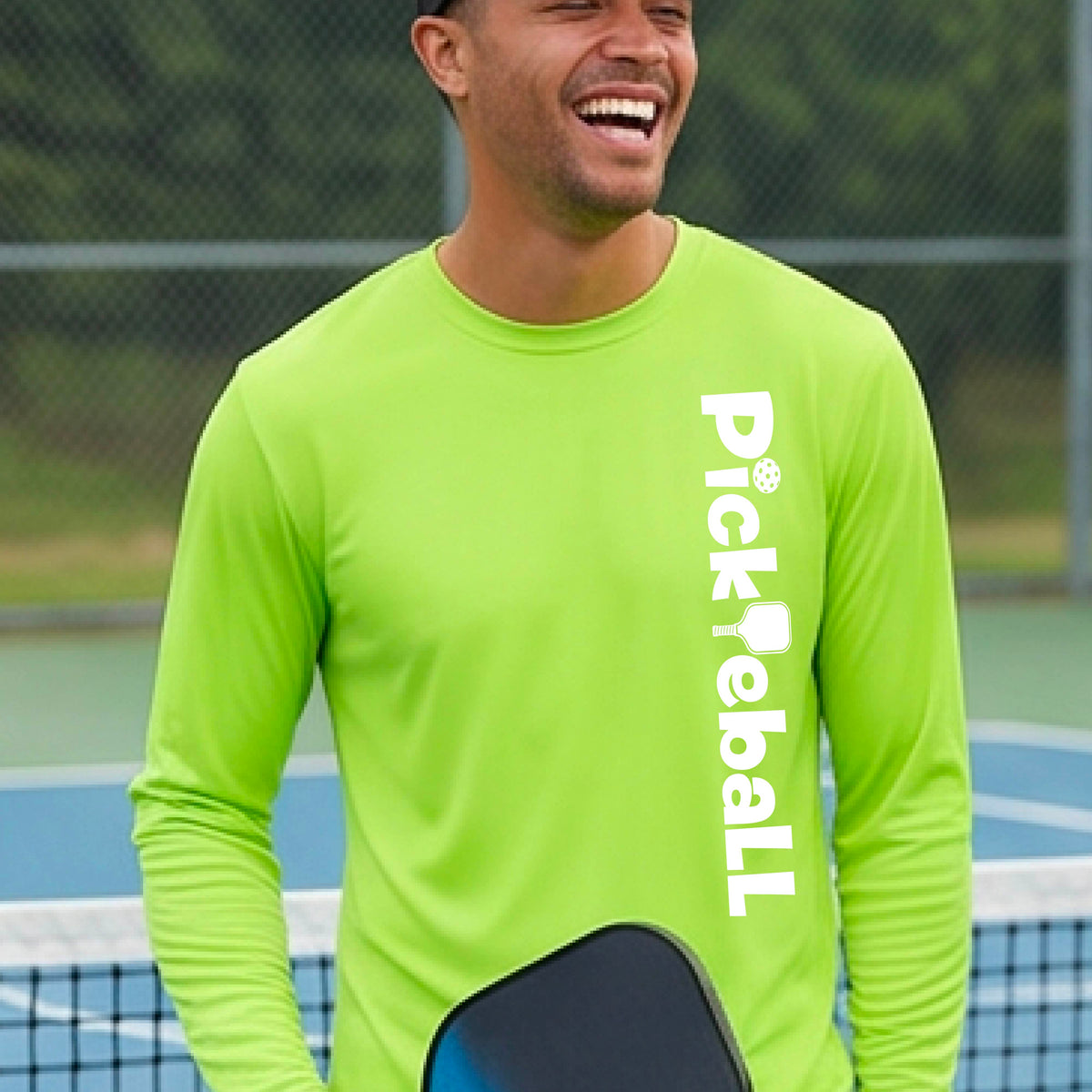 A man laughing on a pickleball court wearing a bright lime green long-sleeve shirt with 'Pickleball' printed vertically in white on the right side of the chest.