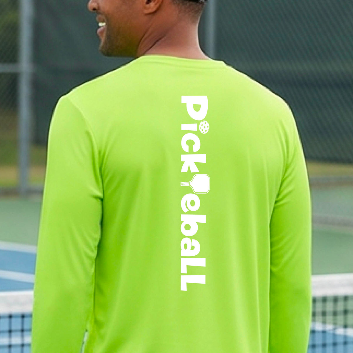 A man viewed from behind wearing a bright lime green long-sleeve athletic shirt with 'Pickball' printed vertically in white on the back, standing on a pickleball court.