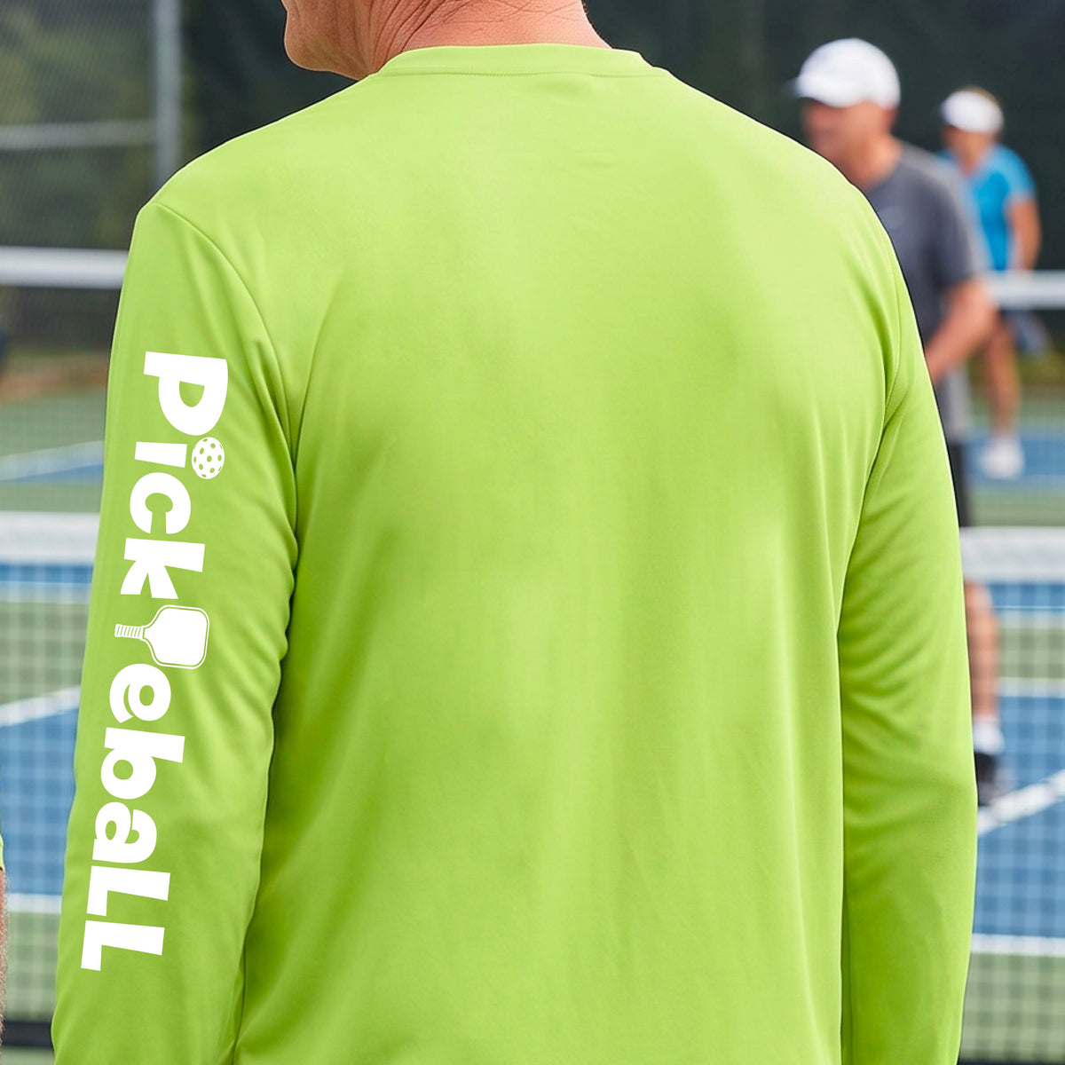 A man viewed from behind wearing a bright lime green long-sleeve athletic shirt with 'Pickleball' printed vertically in white on the left sleeve, standing on a pickleball court with other players blurred in the background.