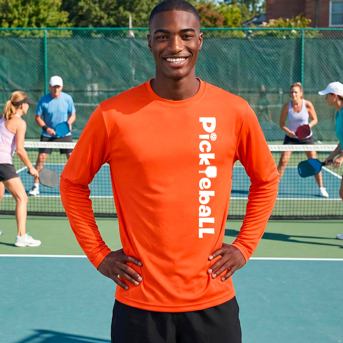 A man smiling on a pickleball court wearing a bright orange long-sleeve athletic shirt with 'Pickleball' printed vertically in white on the front, paired with black shorts.