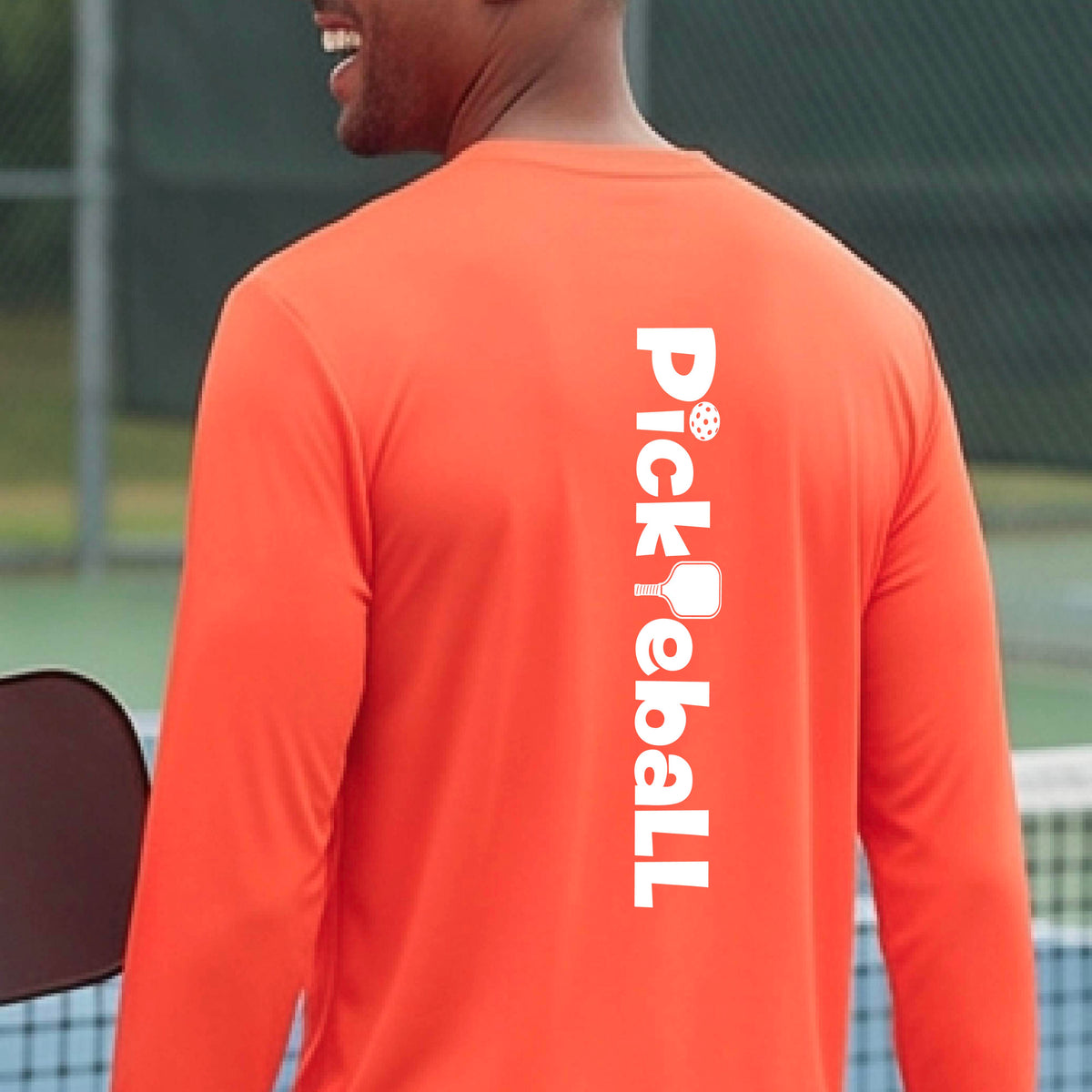 A man wearing a bright orange long-sleeve athletic shirt, viewed from the back. The shirt features the word 'Pickleball' printed vertically in white on the back, with a pickleball icon integrated into the lettering.