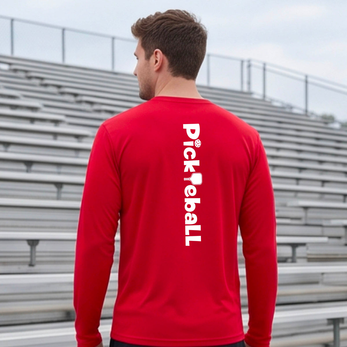 A man viewed from behind wearing a bright red long-sleeve athletic shirt with 'Pickleball' printed vertically in white on the back, standing in front of outdoor stadium bleachers.