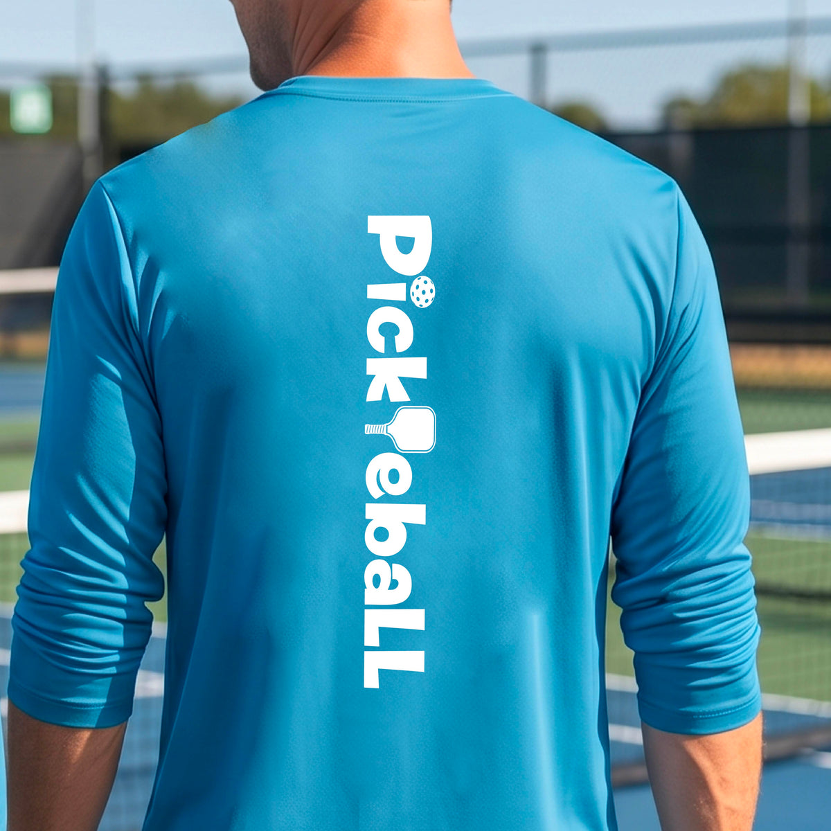 A man viewed from behind wearing a bright blue long-sleeve athletic shirt with 'Pickleball' printed vertically in white on the back, standing on an outdoor pickleball court.