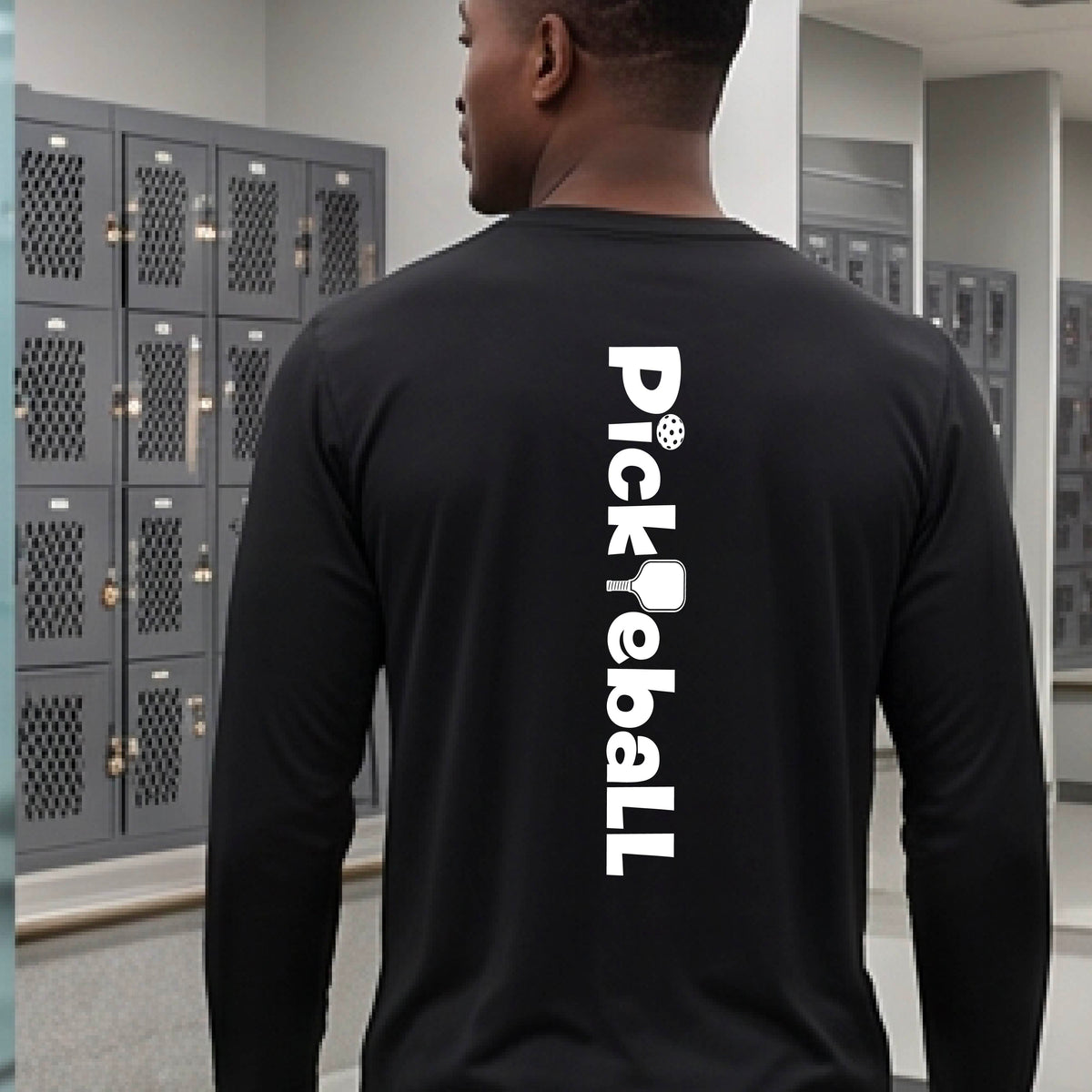 A man viewed from behind wearing a black long-sleeve shirt with a large white vertical 'Pickleball' graphic printed on the back, standing in front of gray school lockers.