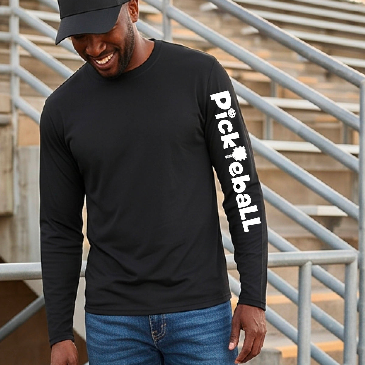 A man wearing a black long-sleeve athletic shirt with 'Pickleball' printed vertically in white on the left sleeve, paired with blue jeans and a black cap, standing in front of outdoor stadium bleachers.