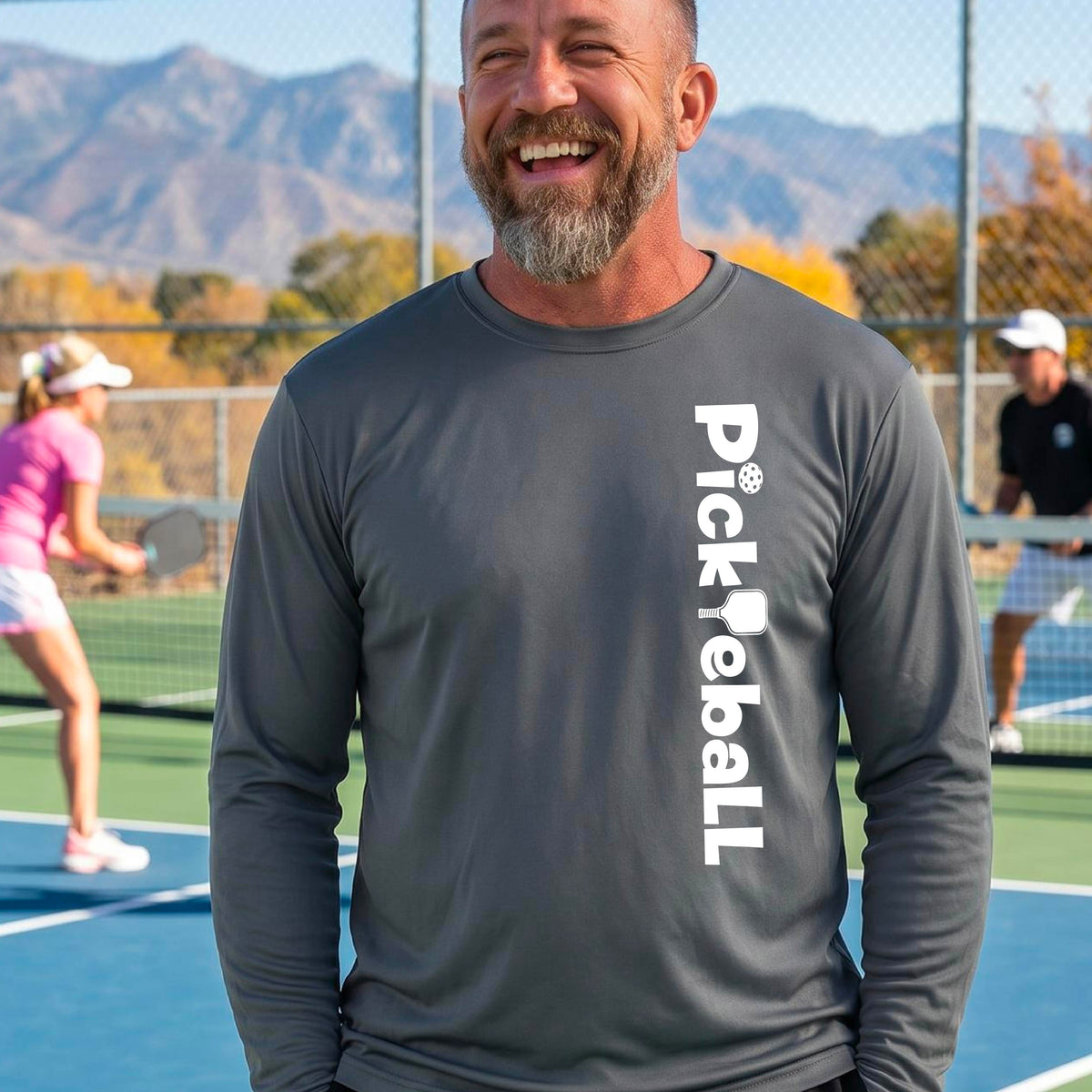 A smiling bearded man wearing a dark gray long-sleeve t-shirt with 'PICKLEBALL' printed vertically in white on the front, standing on an outdoor pickleball court with mountains and other players in the background.
