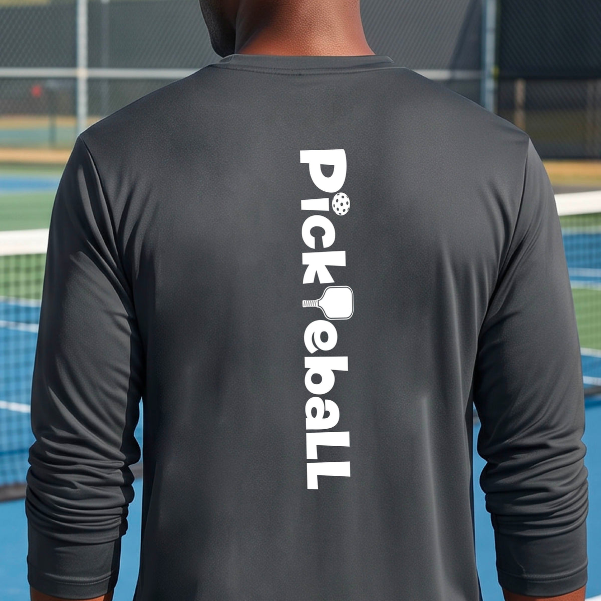 A man viewed from behind wearing a dark gray long-sleeve athletic shirt with 'Pickleball' printed vertically in white on the back, standing on a blue and green pickleball court.