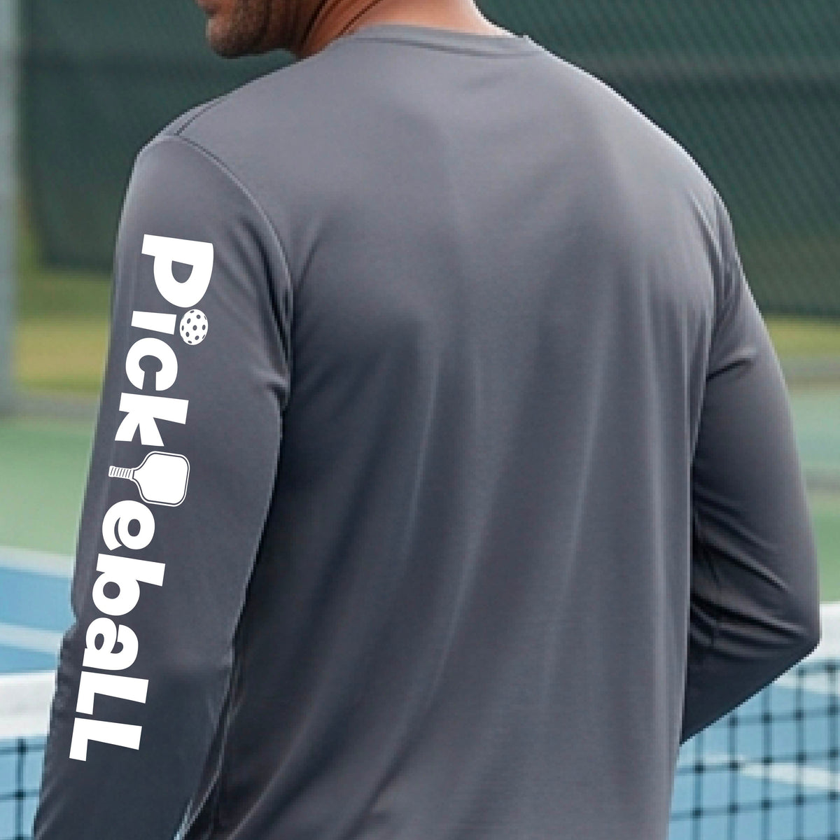 A man wearing a dark gray long-sleeve athletic shirt with 'Pickball' printed in white down the left sleeve, standing on a tennis court.