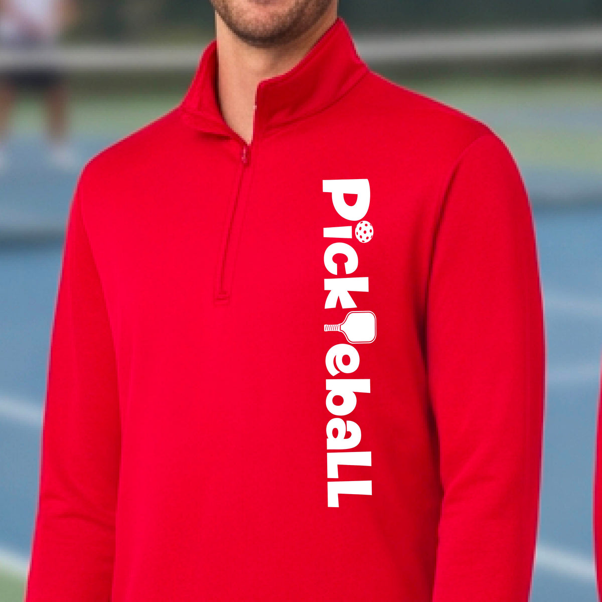 A man wearing a red quarter-zip shirt with a white graphic text 'PICKLEBALL' with a pickleball paddle replacing the L, on the front, standing on a pickleball court.