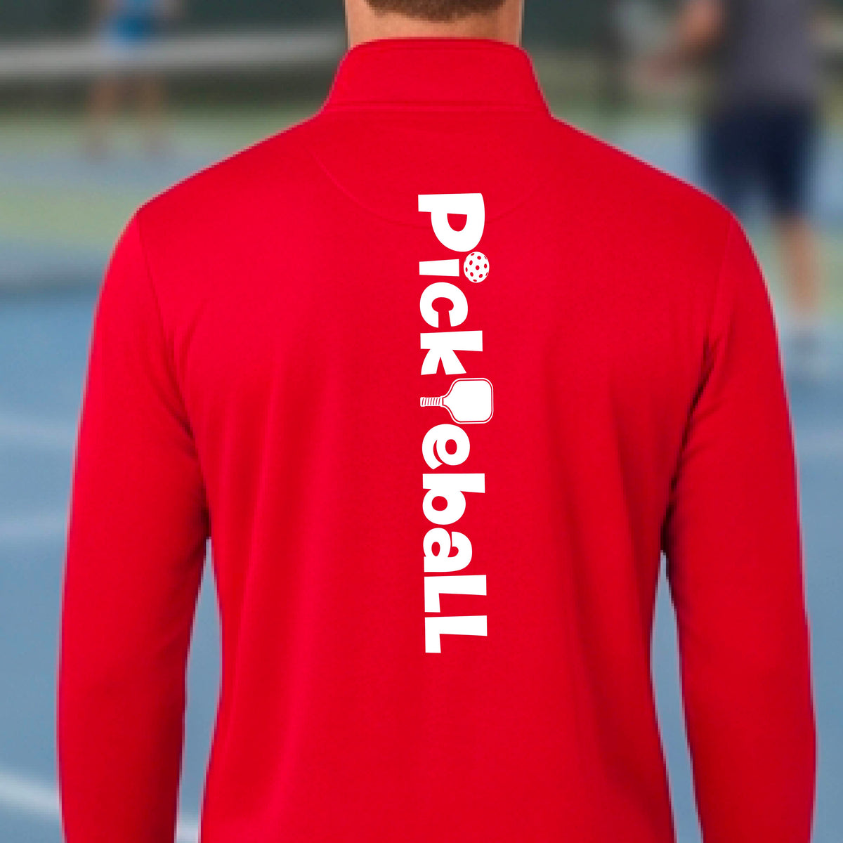 A man wearing a royal blue quarter-zip shirt with a white graphic text 'PICKLEBALL' with a pickleball paddle replacing the L, on the back, standing on a pickleball court.