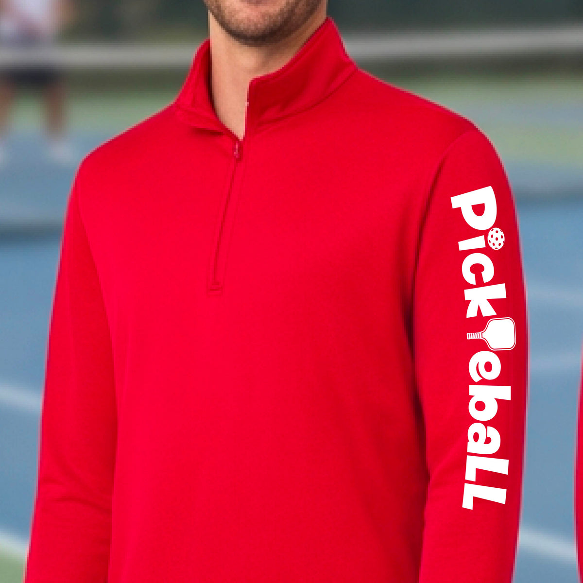 A man wearing a bright red long-sleeve quarter-zip pullover with 'Pickleball' printed vertically in white on the right sleeve, set against a blurred blue pickleball court background.