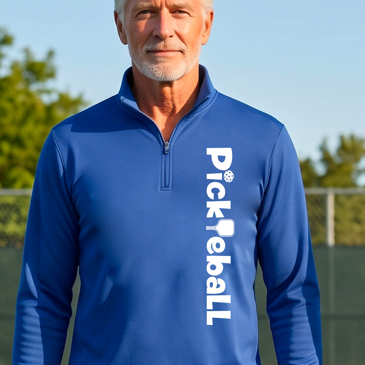 A man wearing a royal blue quarter-zip shirt with a white graphic text 'PICKLEBALL' with a pickleball paddle replacing the L, on the front, standing on a pickleball court.