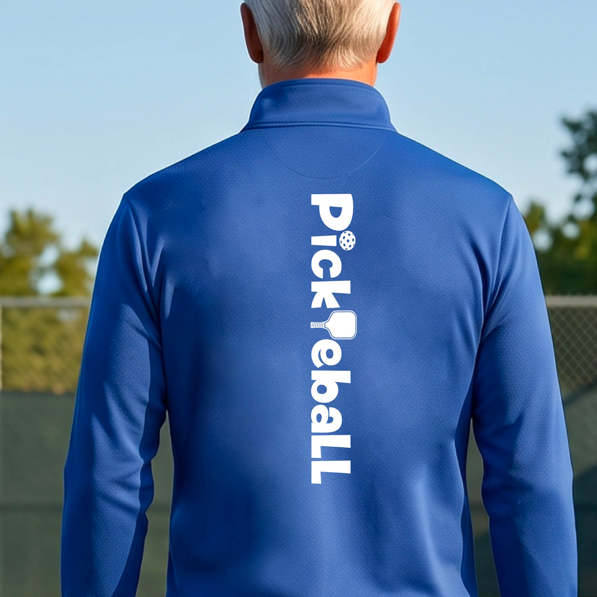 A man viewed from behind wearing a royal blue athletic quarter-zip jacket with 'Pickball' printed vertically in white on the back, set against an outdoor background of a pickleball court and sky.
