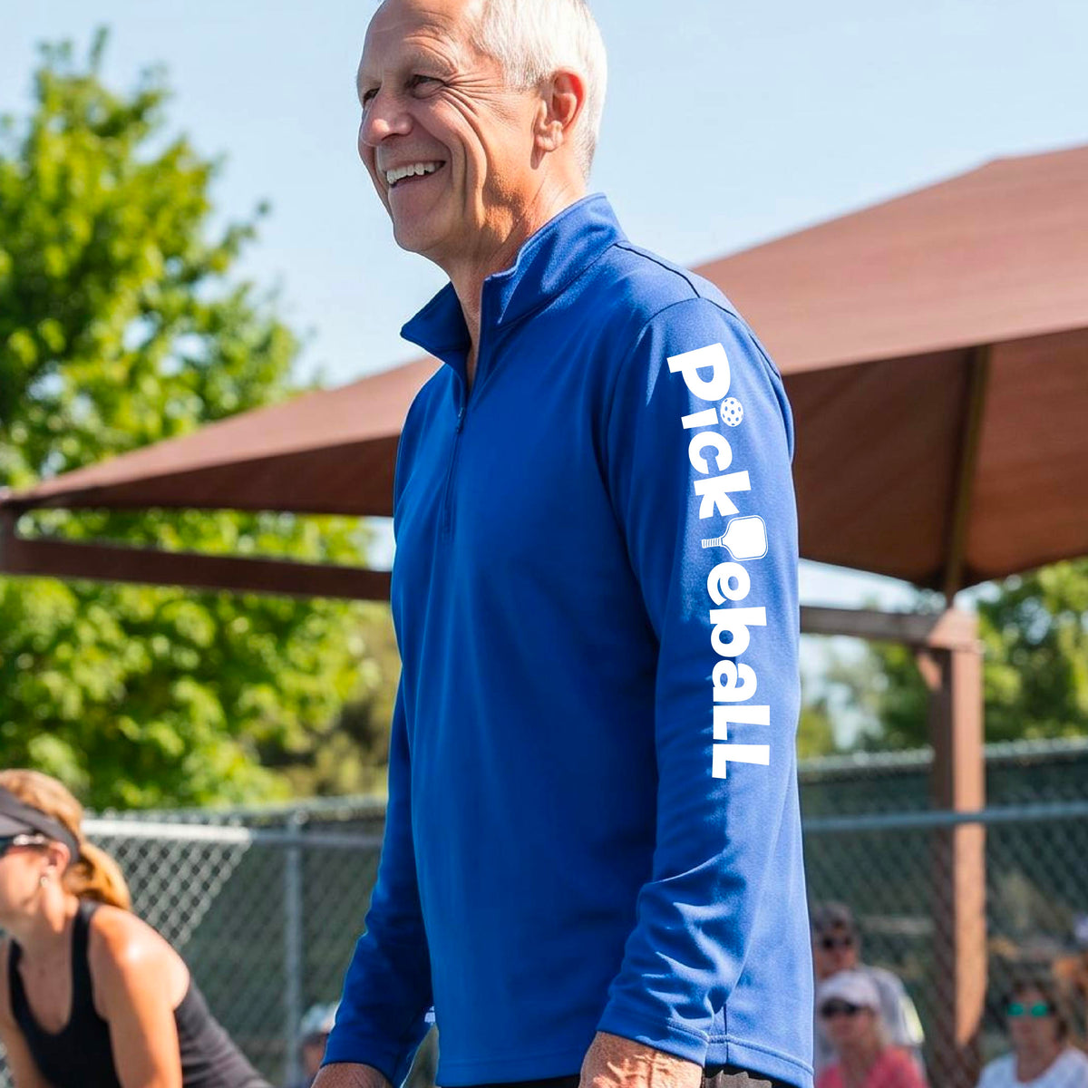 A man wearing a bright royal blue quarter-zip pullover with 'Pickleball' printed vertically in white on the sleeve, standing outdoors on a pickleball court under a brown canopy.