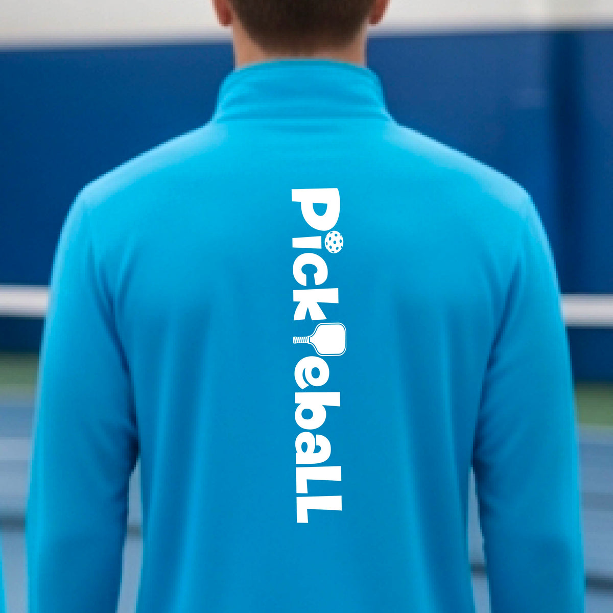 A person viewed from behind wearing a bright blue athletic quarter-zip jacket with the word 'Pickleball' printed vertically in white on the back, standing on a blue pickleball court.