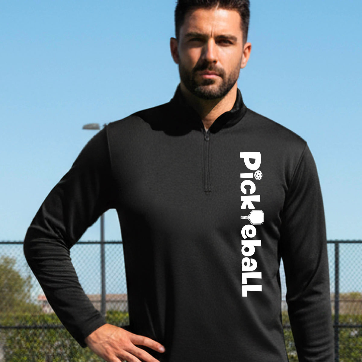 A man wearing a black long-sleeve quarter-zip pullover with 'Pickleball' printed vertically in white on the left side, standing outdoors against a blue sky and fence background.