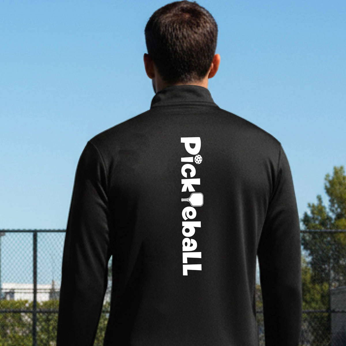 A man wearing a blackquarter-zip shirt with a white graphic text 'PICKLEBALL' with a pickleball paddle replacing the L, on the back, standing on a pickleball court.