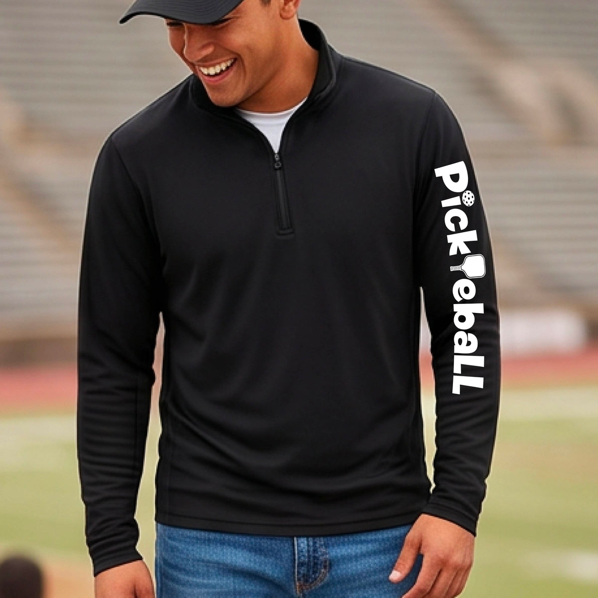 A man wearing a black long-sleeve quarter-zip performance pullover with 'Pickleball' printed vertically in white on the right sleeve, paired with blue jeans and a black cap, set against a blurred stadium background.