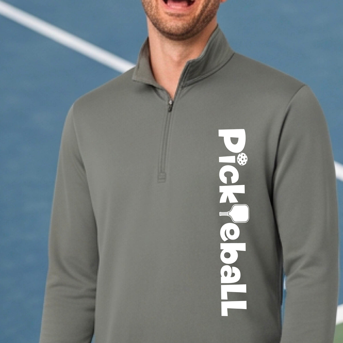 A man wearing a gray quarter-zip pullover with 'Pickleball' printed vertically in white on the right side of the chest, set against a blue pickelball court background.