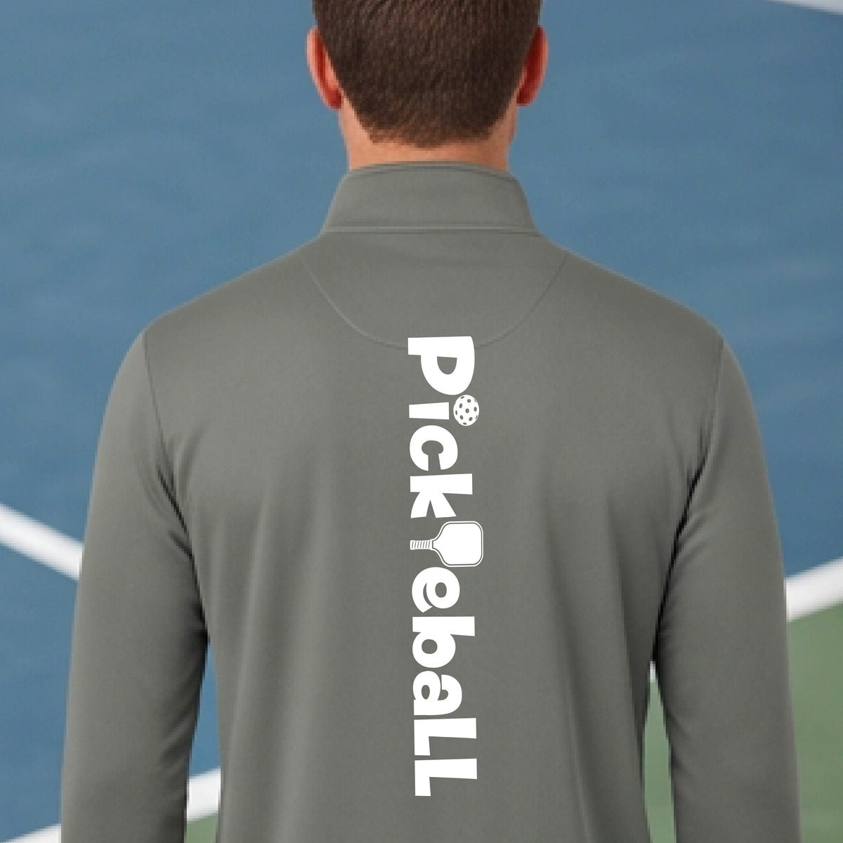 A man viewed from behind wearing a gray long-sleeve athletic quarter-zip pullover with 'Pickleball' printed vertically in white on the back, set against a blue pickleball court background.