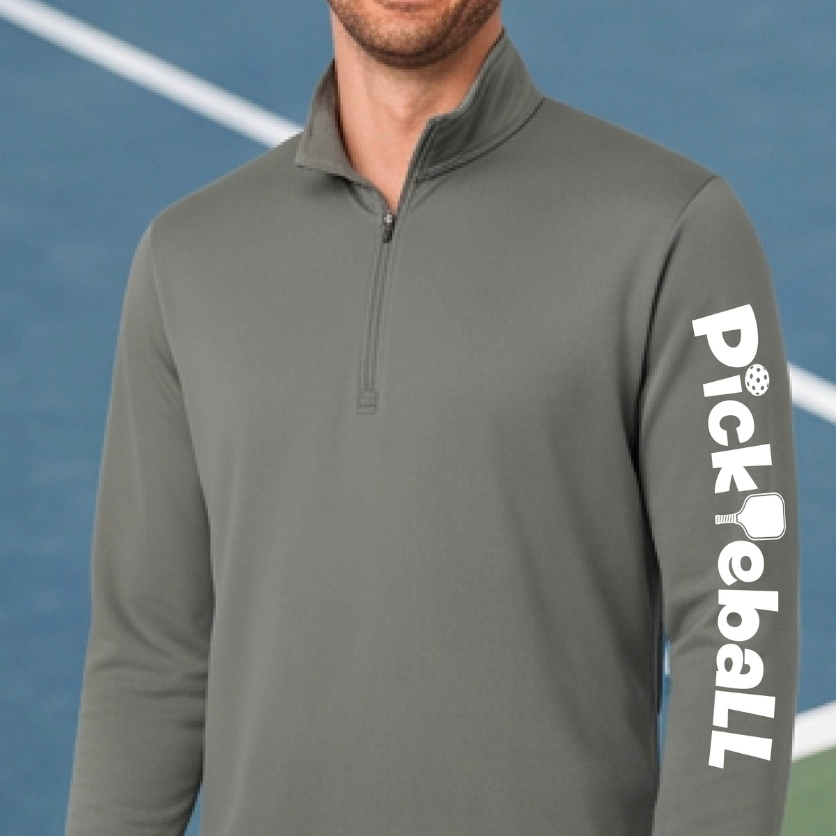 A man wearing a gray quarter-zip long-sleeve shirt with 'Pickleball' printed in white down the right sleeve, set against a blue pickleball court background.