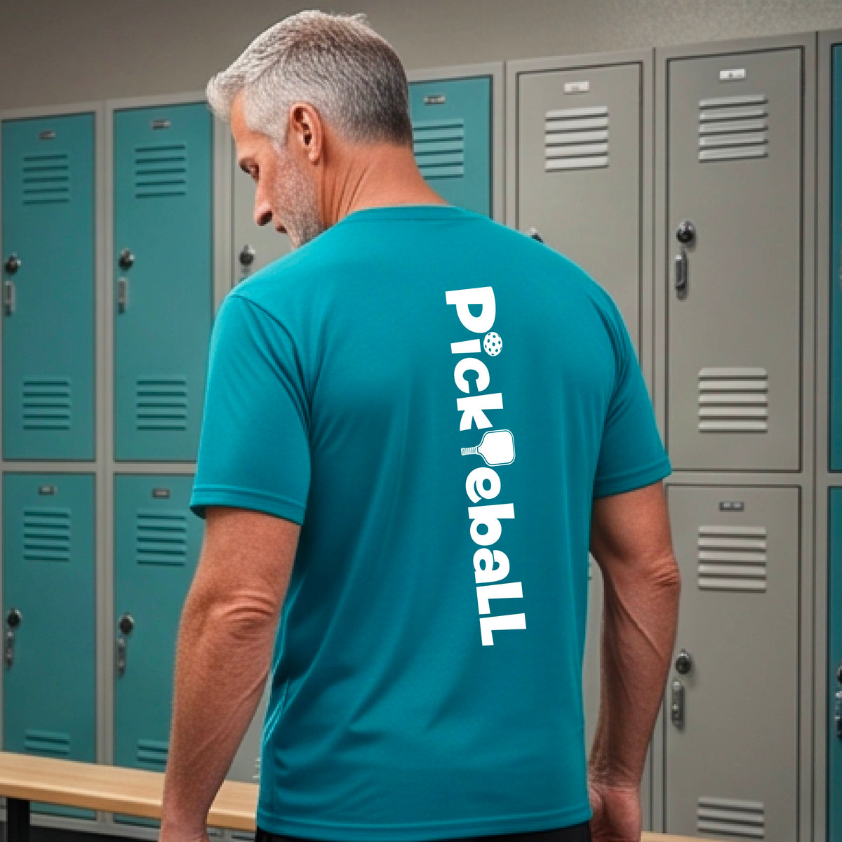 A man wearing a teal short-sleeve t-shirt with a white graphic text 'PICKLEBALL' with a pickleball paddle replacing the L, on the back, standing on a pickleball court.