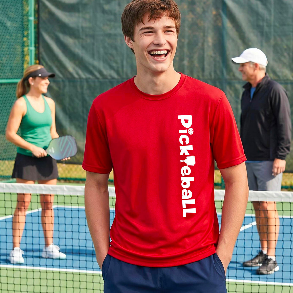 A man wearing a red short-sleeve t-shirt with a white graphic text 'PICKLEBALL' with a pickleball paddle replacing the L, on the front, standing on a pickleball court.