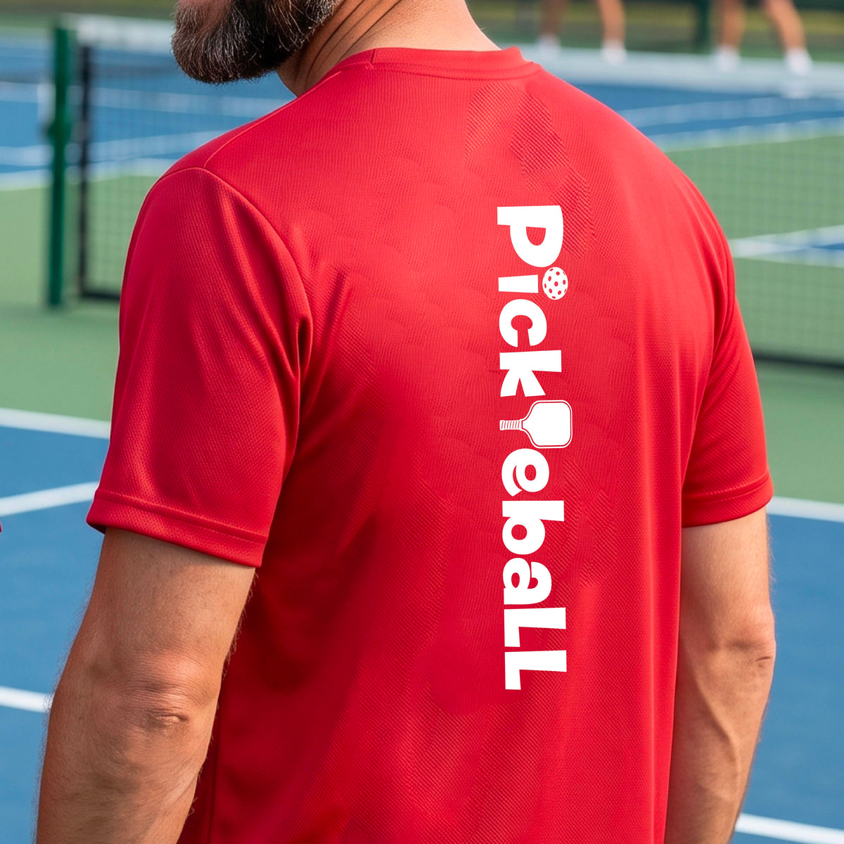 A man wearing a red short-sleeve t-shirt with a white graphic text 'PICKLEBALL' with a pickleball paddle replacing the L, on the back, standing on a pickleball court.