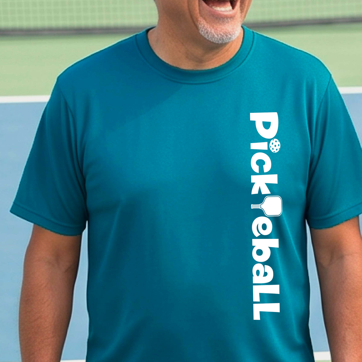 A man wearing a teal short-sleeve t-shirt with a white graphic text 'PICKLEBALL' with a pickleball paddle replacing the L, on the front, standing on a pickleball court.