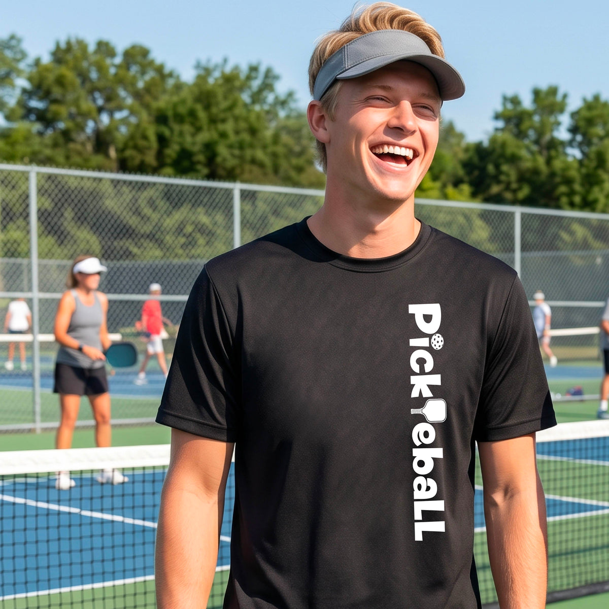 A man wearing a black short-sleeve t-shirt with a white graphic text 'PICKLEBALL' with a pickleball paddle replacing the L, on the front, standing on a pickleball court.