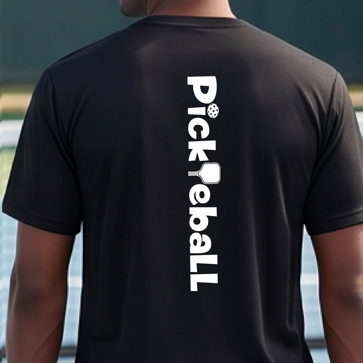 A man wearing a black short-sleeve t-shirt with a white graphic text 'PICKLEBALL' with a pickleball paddle replacing the L, on the back, standing on a pickleball court.