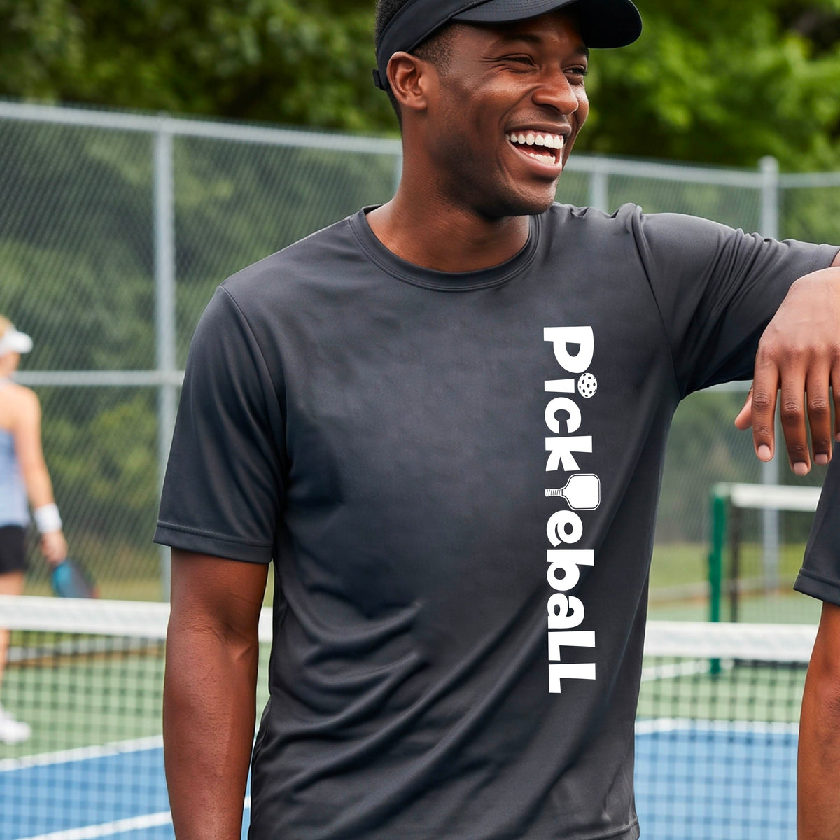 A man wearing a gray short-sleeve t-shirt with a white graphic text 'PICKLEBALL' with a pickleball paddle replacing the L, on the front, standing on a pickleball court.
