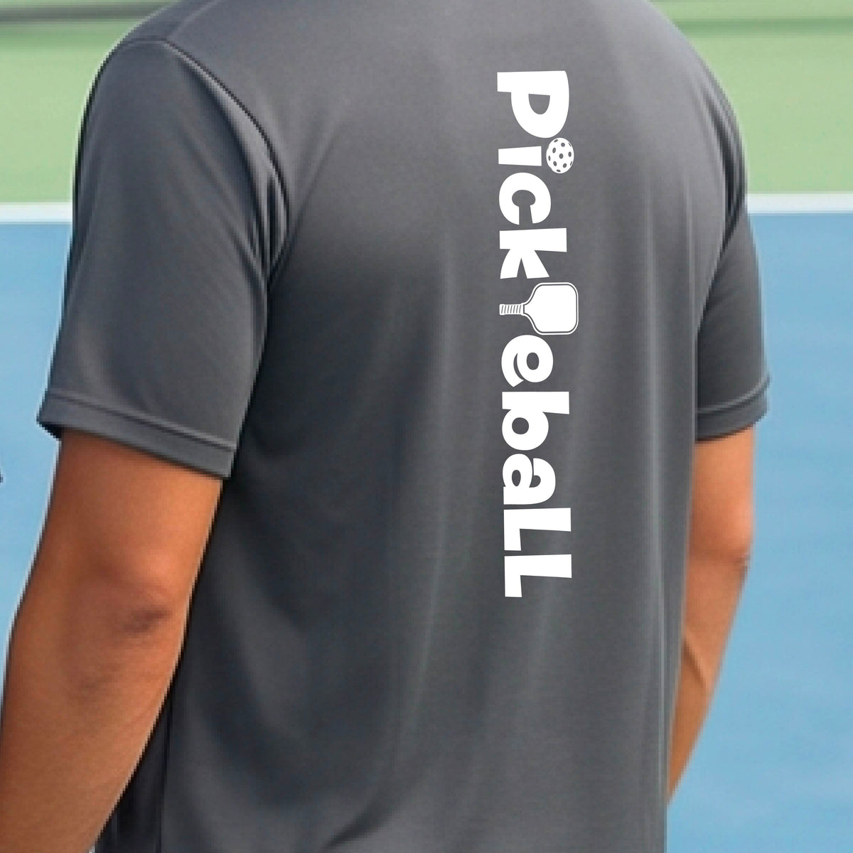 A man wearing a gray short-sleeve t-shirt with a white graphic text 'PICKLEBALL' with a pickleball paddle replacing the L, on the back, standing on a pickleball court.