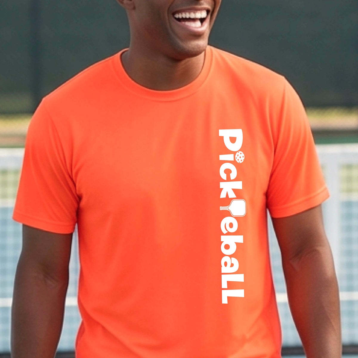A man wearing an orange short-sleeve t-shirt with a white graphic text 'PICKLEBALL' with a pickleball paddle replacing the L, on the front, standing on a pickleball court.
