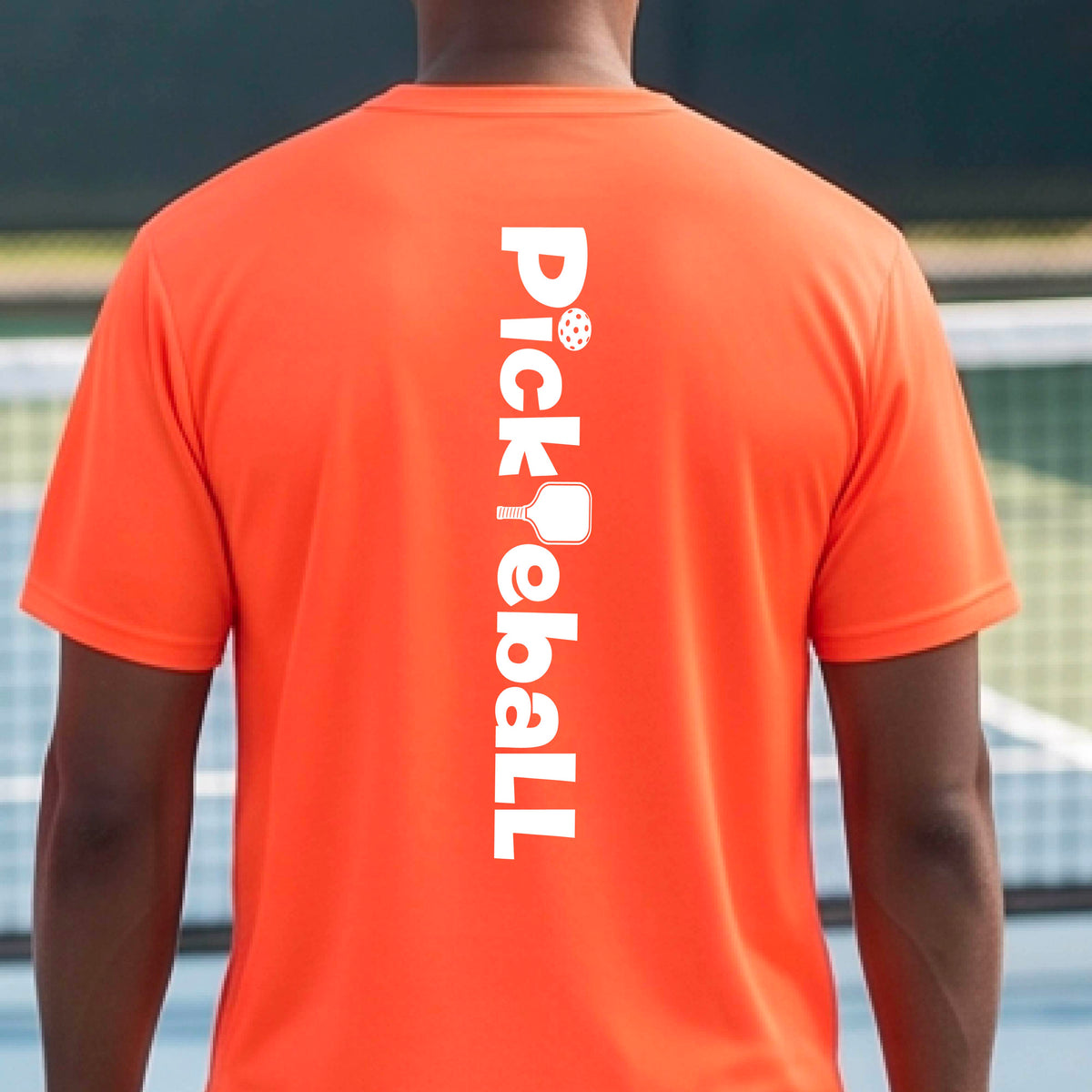 A man wearing an orange short-sleeve t-shirt with a white graphic text 'PICKLEBALL' with a pickleball paddle replacing the L, on the back, standing on a pickleball court.