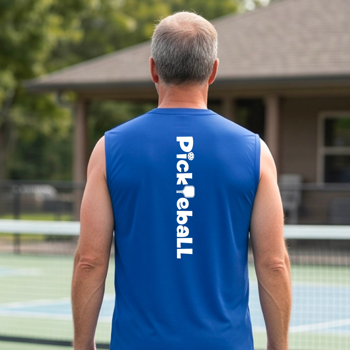 A man wearing a royal blue sleeveless t-shirt with a white graphic text 'PICKLEBALL' with a pickleball paddle replacing the L, on the back, standing on a pickleball court.